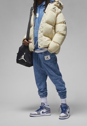 Person iført en cremefarvet dunjakke, blå hættetrøje, blå joggingbukser, hvide høje sneakers og holder en sort taske med hvidt logo.