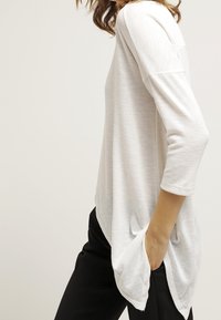 Weiße, langärmlige Bluse mit lockerer Passform, abgerundetem Saum und seitlichen Taschen. Der Stoff wirkt weich mit einer leichten Textur.