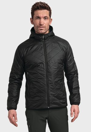 Mann trägt schwarze, gesteppte Reißverschlussjacke mit Kapuze und schwarze Hose, steht vor einfarbigem hellem Hintergrund.