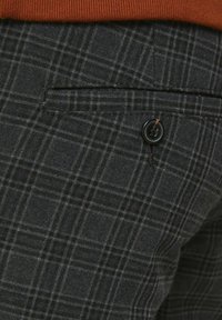 Gros plan sur un pantalon à carreaux gris foncé avec une poche arrière noire boutonnée, associé à un pull orange rouille.