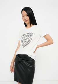 Giovane donna con lunghi capelli neri che indossa una maglietta bianca con un design di tigre e una gonna di pelle nera con dettaglio a fiocco.