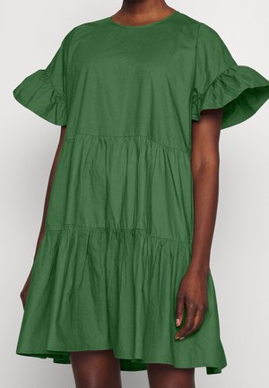 Femme portant une robe verte ample à manches courtes avec des volants aux manches et une jupe à étages, debout devant un fond uni.