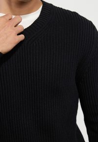 Granatowy sweter z dzianiny z dekoltem w kształcie litery V, z prążkowaną teksturą i długimi rękawami. Zbliżenie na dekolt i ramię.