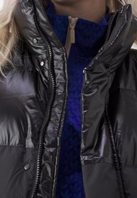 Schwarze Pufferjacke mit glänzender Textur, ausgestattet mit einem Reißverschluss vorne, Druckknöpfen und einem blauen gemusterten Innenfutter.