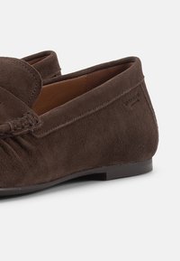 Καφέ μοκασίνι από σουέντ με ραμμένο σχεδιασμό moc toe και χαμηλή μαύρη σόλα, με λεπτομέρειες από το πλάι και από πάνω σε λευκό φόντο.