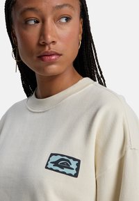 Μπεζ φούτερ με ριμπ λαιμόκοψη, με ορθογώνιο μπλε και μαύρο λογότυπο στο στήθος. Λεία υφή, casual σχέδιο.