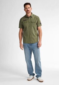 Chemise à manches courtes vert olive avec poches frontales, fermeture par boutons et écussons aux épaules, associée à un jean bleu clair et des baskets blanches.
