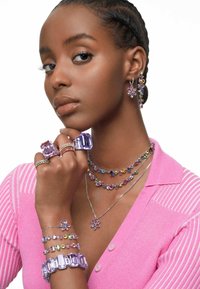 Purple and multicolored jewelry, including rings, earrings, and layered necklaces, set against a plain background. Each piece features sparkling gemstones.