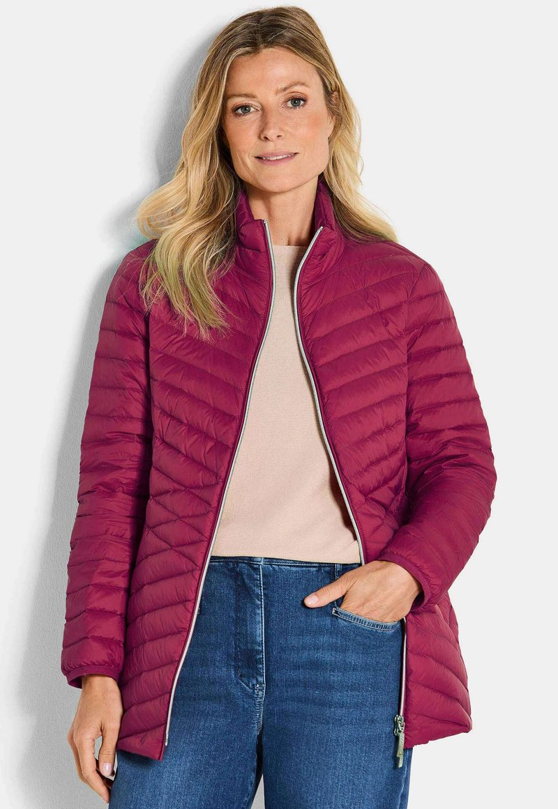 Vrouw in een geborduurde burgundy jack met rits over een beige top en blauwe jeans, staand tegen een witte achtergrond.