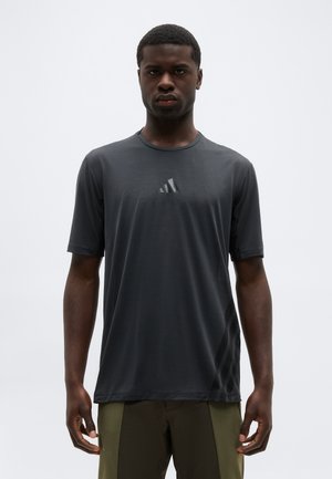 Muž oblečený v tmavosivej krátkosleevej košeli Adidas a olivovo zelených nohaviciach stojaci pred jednoduchým svetlým pozadím.