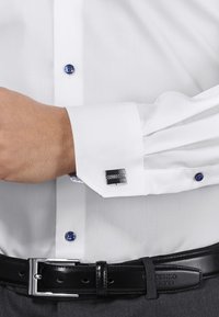 Witte overhemd met blauwe knoop en manchetknopen. Manchetknop heeft een gestructureerd zilveren oppervlak. Zwarte leren riem met zilveren gesp.