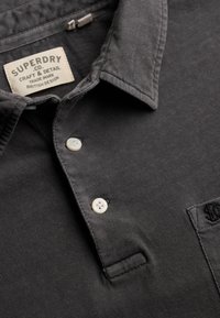 Col de chemise Superdry gris foncé à boutons crème avec étiquette de marque visible cousue à l'intérieur de l'encolure.