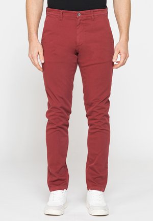 Uomo con pantaloni slim fit rosso scuro e sneakers bianche, in piedi davanti a uno sfondo bianco uniforme.