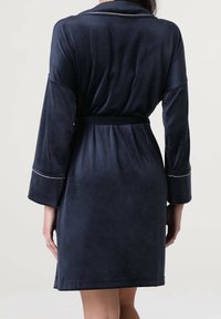 Donna che indossa una veste di velluto blu scuro, lunga fino al ginocchio, con maniche lunghe e una cintura in vita, mostrata di spalle su uno sfondo neutro.