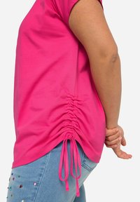 Camiseta de algodón rosa con detalle lateral fruncido y lazos ajustables; mangas cortas; combinada con jeans de mezclilla azul claro con embellishments.