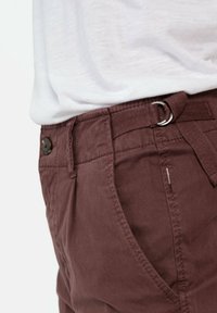 Gros plan sur un pantalon marron avec poche latérale, fermeture par bouton et ceinture en tissu ajustable avec anneau métallique, porté avec une chemise blanche.
