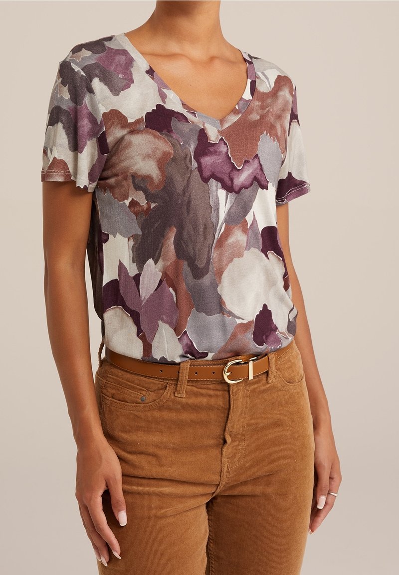 Korte mouwen, V-hals top in een multicolore bloemenpatroon met tinten paars, kastanjebruin en beige, gecombineerd met bruine corduroy broek en een beige riem.