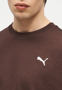 Νεαρό άτομο που φοράει καφέ πουκάμισο με λευκό λογότυπο Puma στο αριστερό στήθος, ορατό από το πηγούνι μέχρι το πάνω μέρος του κορμού.