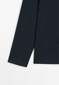 Langer Ärmel eines schwarzen Kleidungsstücks mit sichtbaren Nähten an Bündchen und Saum vor einem weißen Hintergrund.