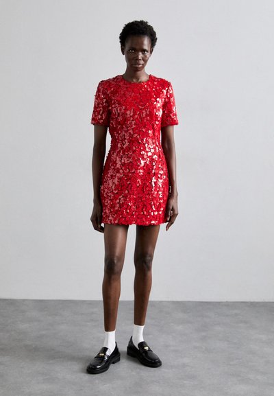 Robe en sequins rouges avec des manches courtes, un encolure ronde et une silhouette ajustée. Portée avec des mocassins noirs et des chaussettes blanches. Fond uni.