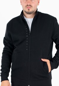 Czarny rozpinany sweatshirt z wysokim kołnierzem, ściśle dopasowanymi mankietami i dołem, z subtelnym logo na froncie. Miękka tkanina o gładkiej fakturze.