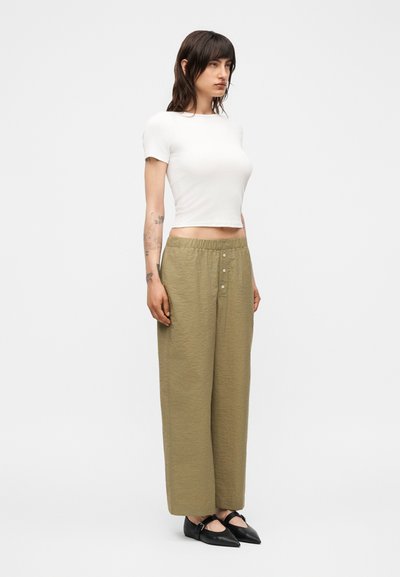 Γυναίκα με σκούρα μαλλιά που φοράει εφαρμοστό λευκό κοντομάνικο crop top, άνετο ελαιόχρωμο παντελόνι και μαύρα μυτερά μπαλαρίνια, στέκεται μπροστά σε λευκό φόντο.