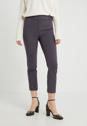 Vrouw draagt donkergrijze, slim fit cropped broek, crèmekleurige trui met lange manchetten en zwarte enkellaarsjes met blokhak en studs.