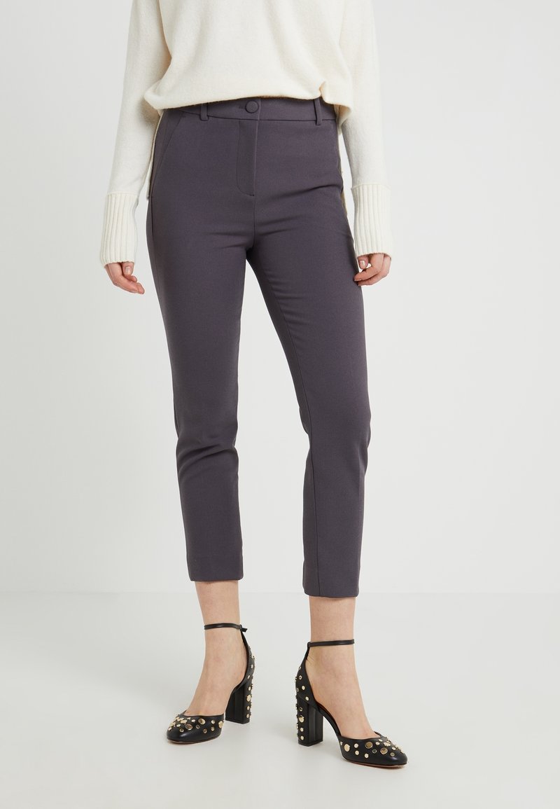 Femme portant un pantalon slim court gris foncé, un pull crème avec de longues poignets, et des chaussures noires à talon bloc, à bride cheville et ornées de clous.