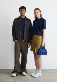 Νεαρός άνδρας με σκούρο μπουφάν και καφέ παντελόνι στέκεται δίπλα σε γυναίκα με μπλε μπλουζάκι, κίτρινη καρό φούστα και λευκά αθλητικά, κρατώντας μπλε τσάντα, πάνω σε γκρι πάτωμα.