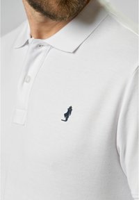 White polo shirt made from textured fabric, featuring a collar and a small navy embroidered figure accent on the left chest.