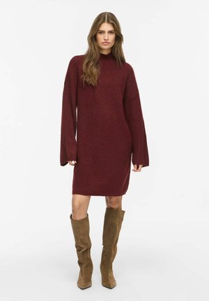 Woman wearing a long-sleeve maroon sweater dress and brown suede knee-high boots standing against a white background.
