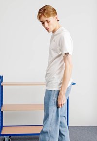 T-shirt de algodão branca com mangas curtas e um logótipo discreto; combinado com jeans de denim de corte solto em azul claro. Prateleiras de madeira ao fundo.