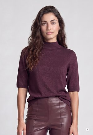 Frau mit langen, welligen Haaren, die ein dunkelviolettes Kurzarm-Top und passende Lederhosen trägt, steht vor einem schlichten hellen Hintergrund.