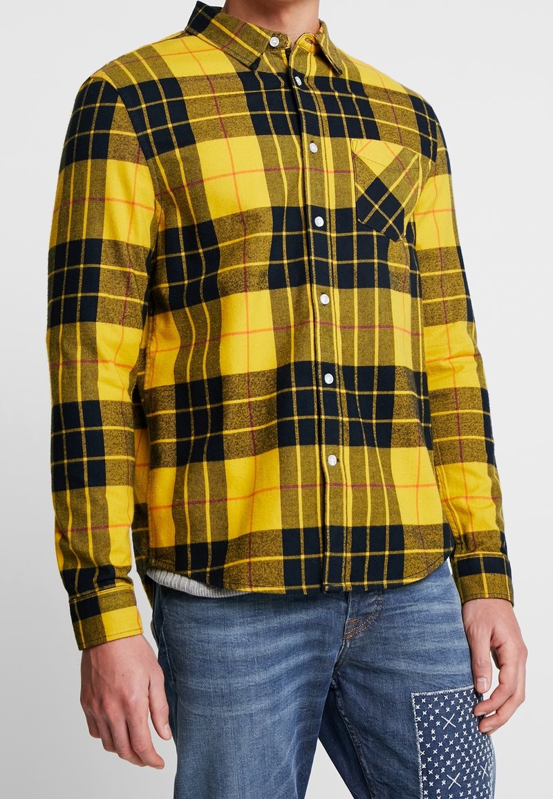 Homme portant une chemise à carreaux jaune et noire boutonnée avec une poche sur la poitrine, associée à un jean bleu doté d'un empiècement à motifs sur la cuisse.
