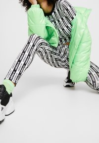 Svartvita mönstrade leggings med "adidas"-text, i kombination med en glänsande ljusgrön puffjacka och svarta träningsskor.