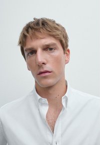 Jeune homme aux cheveux brun clair et aux yeux verts, portant une chemise blanche à boutons sur un fond clair uni.