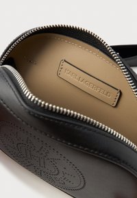 Fekete bőr táska belső rész bézs béléssel, cipzárral záródik és dombornyomott logóval rendelkezik. Egyszerű dizájn, sima textúrával.