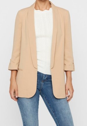 Personne portant un blazer beige aux manches retroussées sur un haut blanc à texture et un jean bleu, debout devant un fond uni.