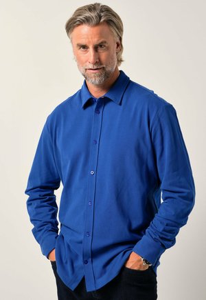 Blå langærmet skjorte lavet af blødt stof, med knaplukning foran, krave og to-farvede knapper. Bæres med mørke jeans.