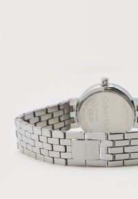 Ρολόι με ασημένιο μεταλλικό βραχιόλι με ορατή σήμανση Calvin Klein στην πίσω πλευρά και στο κλείσιμο, τοποθετημένο πάνω σε λευκή επιφάνεια.