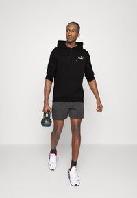 Czarna bluza z kapturem z kieszenią kangurzą, szare szorty i białe sportowe buty z czerwonymi akcentami. Temat trzyma czarny kettlebell.