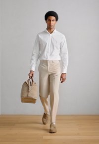 Pantalones beige combinados con una camisa blanca abotonada, sosteniendo un bolso topo con una correa y herrajes de acento, sobre un suelo de madera clara.