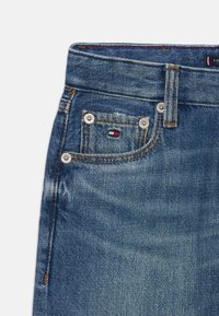 Jeans in denim blu scuro con una texture sbiadita, dotati di una tasca anteriore con hardware argentato e un patch con logo con accenti rossi, bianchi e blu.
