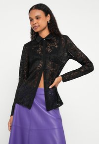 Crna čipkasta košulja s dugim rukavima, kontrastnim crnim ovratnikom i uskim krojem. Nošena preko ljubičaste kožne suknje.