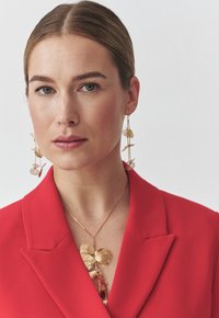 Punane bleiser, millel on teravad kraed, on kantud kuldse avaldustegelase kaelakee kohal, millel on suured lehekujulised aksessuaarid ja tekstuuriga ripats. Julged kõrvarõngad täiendavad välimust.