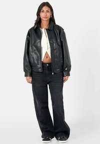 Vrouw in een zwart leren jack, wit cropped overhemd, losse zwarte spijkerbroek en zwarte sneakers, staand tegen een witte achtergrond.