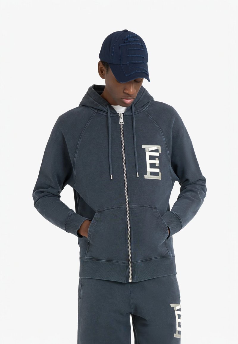 Sweat à capuche zippé bleu marine en tissu doux avec une poche kangourou, présentant un logo bien visible sur la poitrine et des poignets côtelés. Pantalons de jogging visibles.