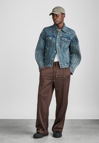 Denim jacket with blue and faded stripes, two front pockets, paired with brown wide-leg pants and a light gray cap. Smooth textures throughout.