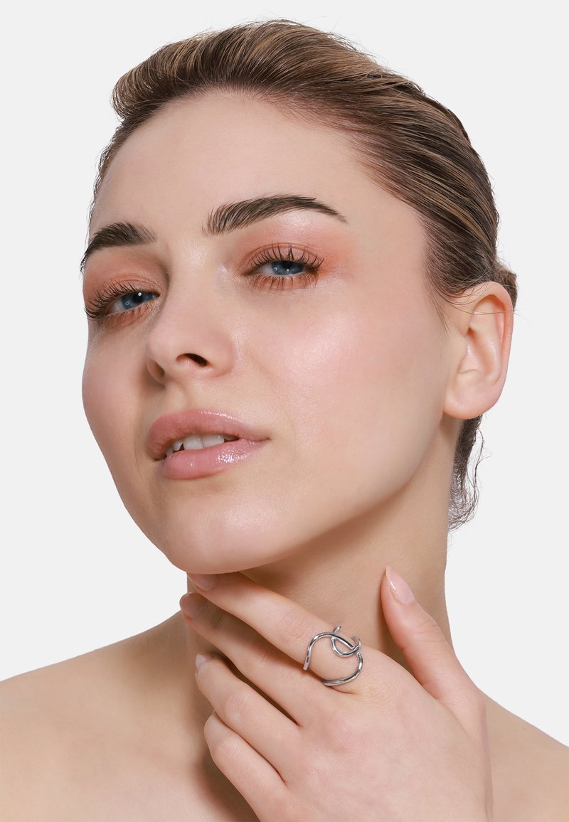 Anello in metallo argento con un design attorcigliato, caratterizzato da curve morbide e una forma aperta, indossato sulla mano di un modello vicino alla linea della mandibola.