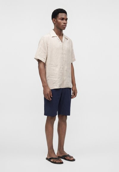 Jeune homme debout, portant une chemise beige à manches courtes boutonnée, un short bleu marine et des tongs noires, sur un fond gris uni.